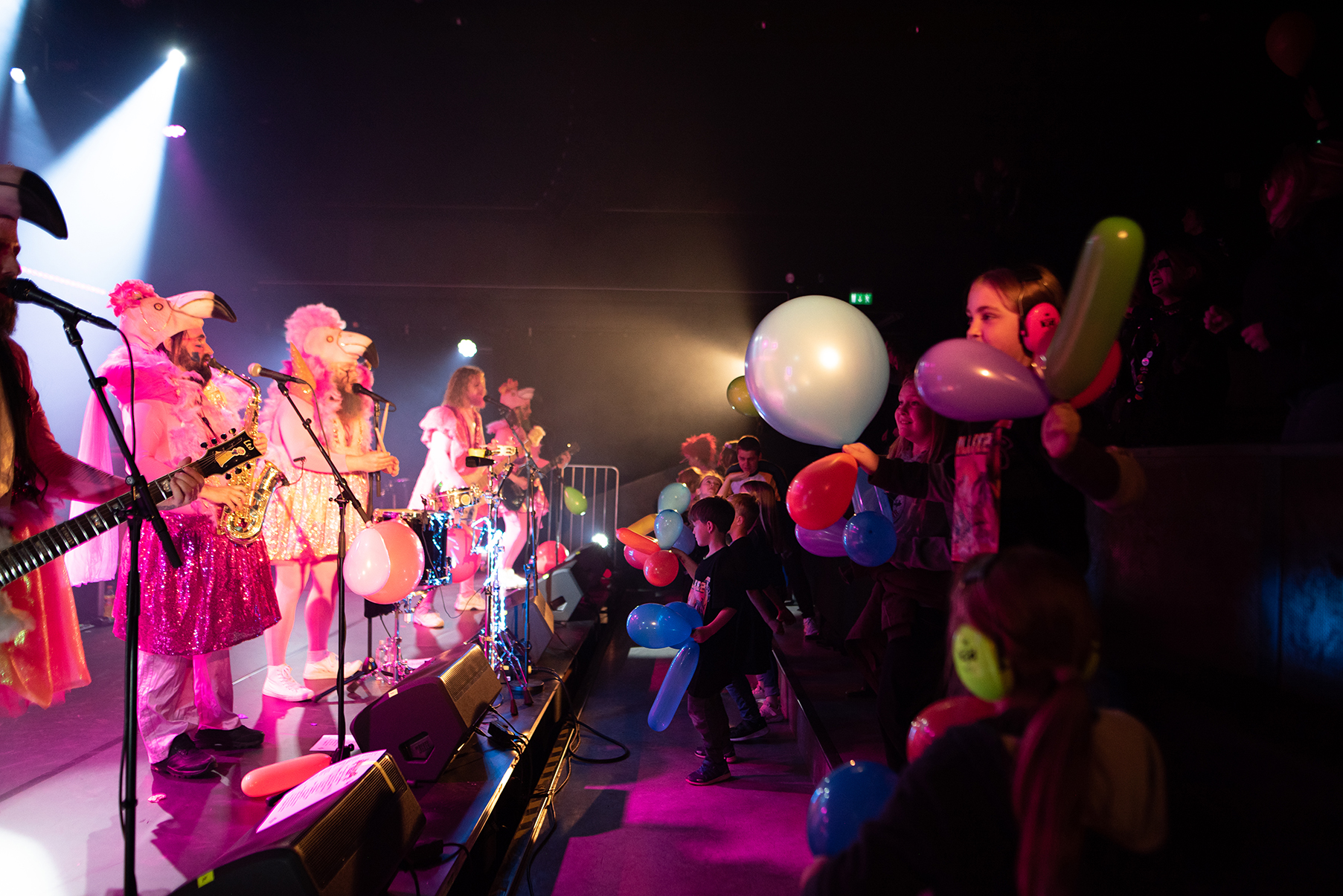 Til venstre en rekke musikere på scenen i et mørkt lokale. Musikerne er ikledd pelikanhatter, rosa og glitrende kostymer. Publikum til høyre består av smilende barn, noen med hørselsvern, som holder fargerike ballonger. 