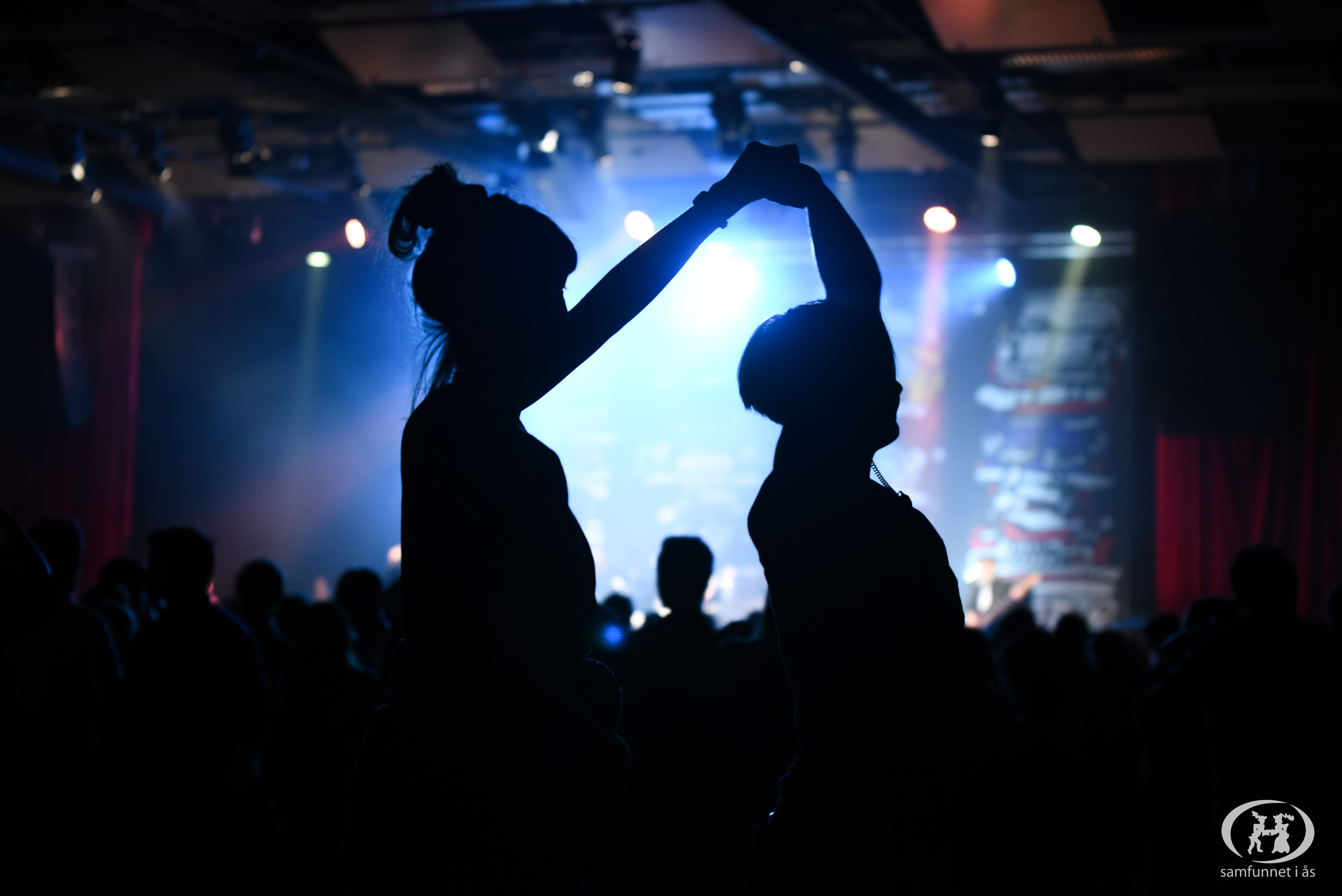 Siluetten av to personer som danser sammen i et mørkt konsertlokale. I bakgrunnen kan man skimte en scenelys og annet publikum. 