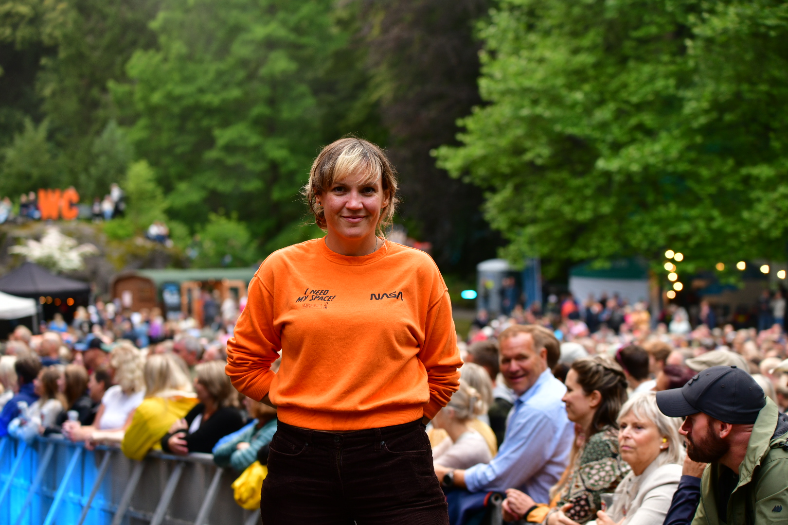 Person ikledd oransje genser på en forhøyning foran barrikadene på en festivalscene. I bakgrunnen et utendørs festivalområde og publikummere. Foto. 