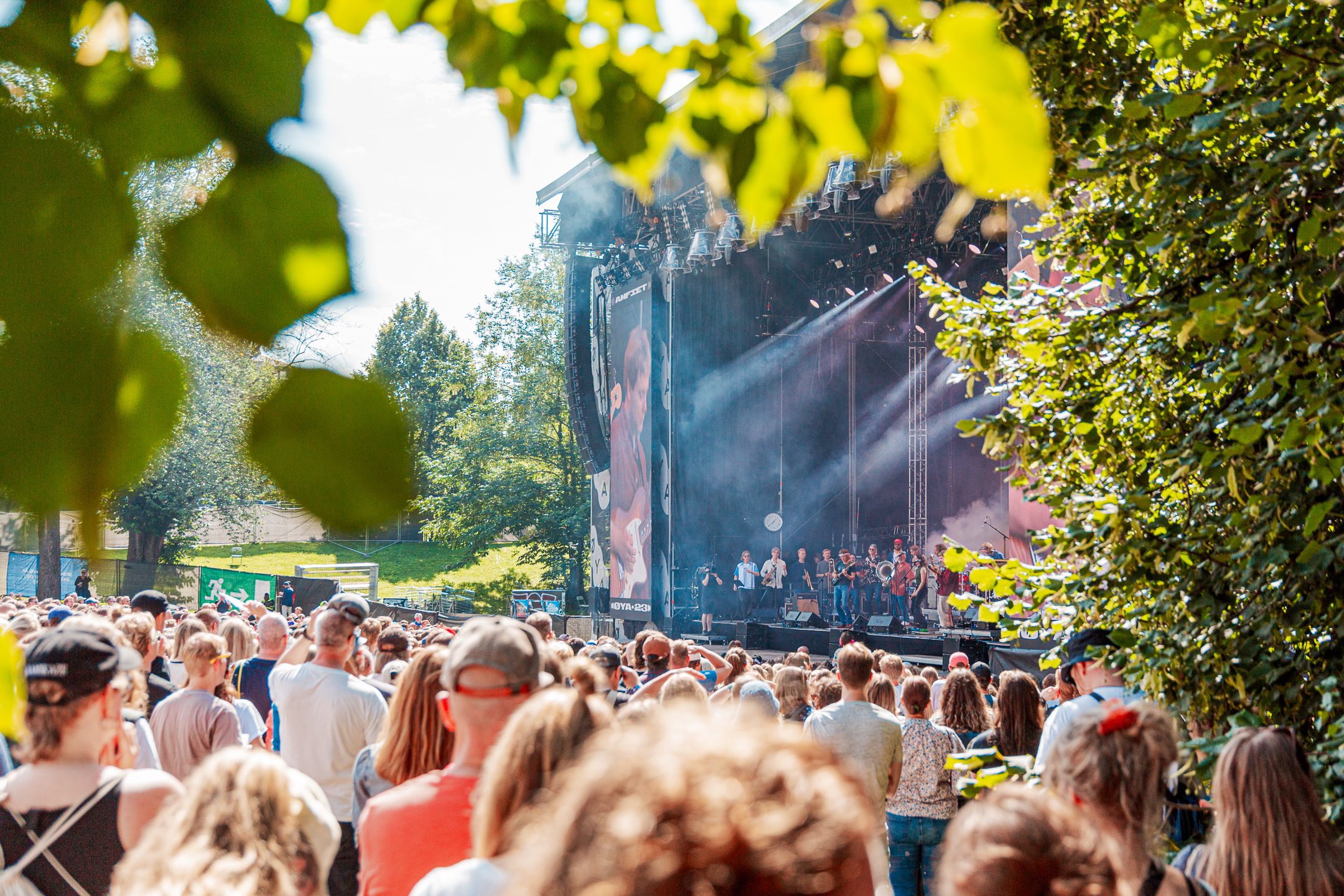Sommer og sol på Øyafestivalen. I solsteken står publikum og nyter en konsert på Amfiet i Tøyenparken. Omringet av grønne trær. På scenen skimtes et stort band med både blåserekke og korister. 