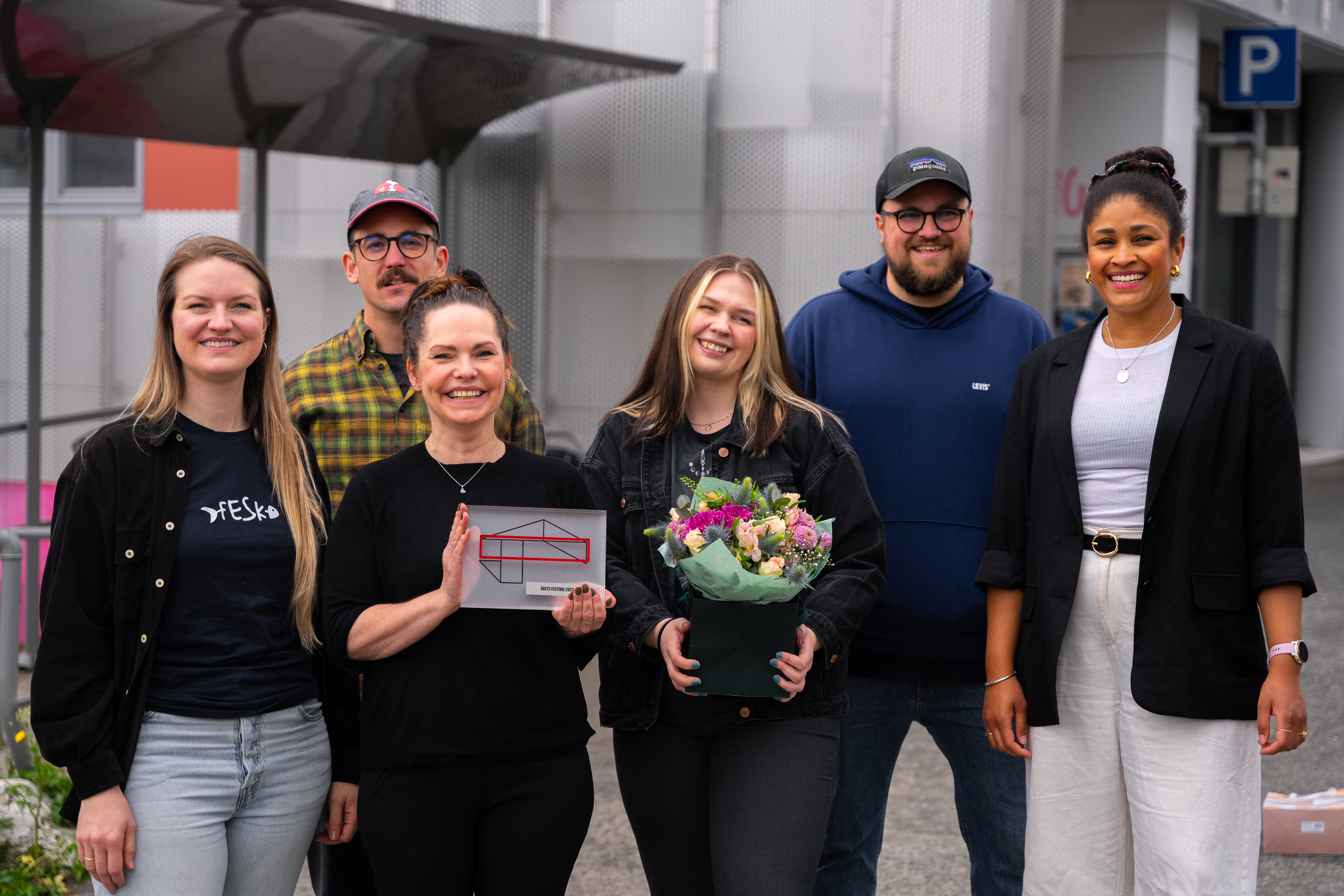 Seks personer oppstilt foran en plakatvegg med festivalplakater smiler fornøyd og tilfreds. Staben i Bukta holder stolt frem pokal og blomster fra Norske Kulturarrangører og festivalplakaten fra 2023.