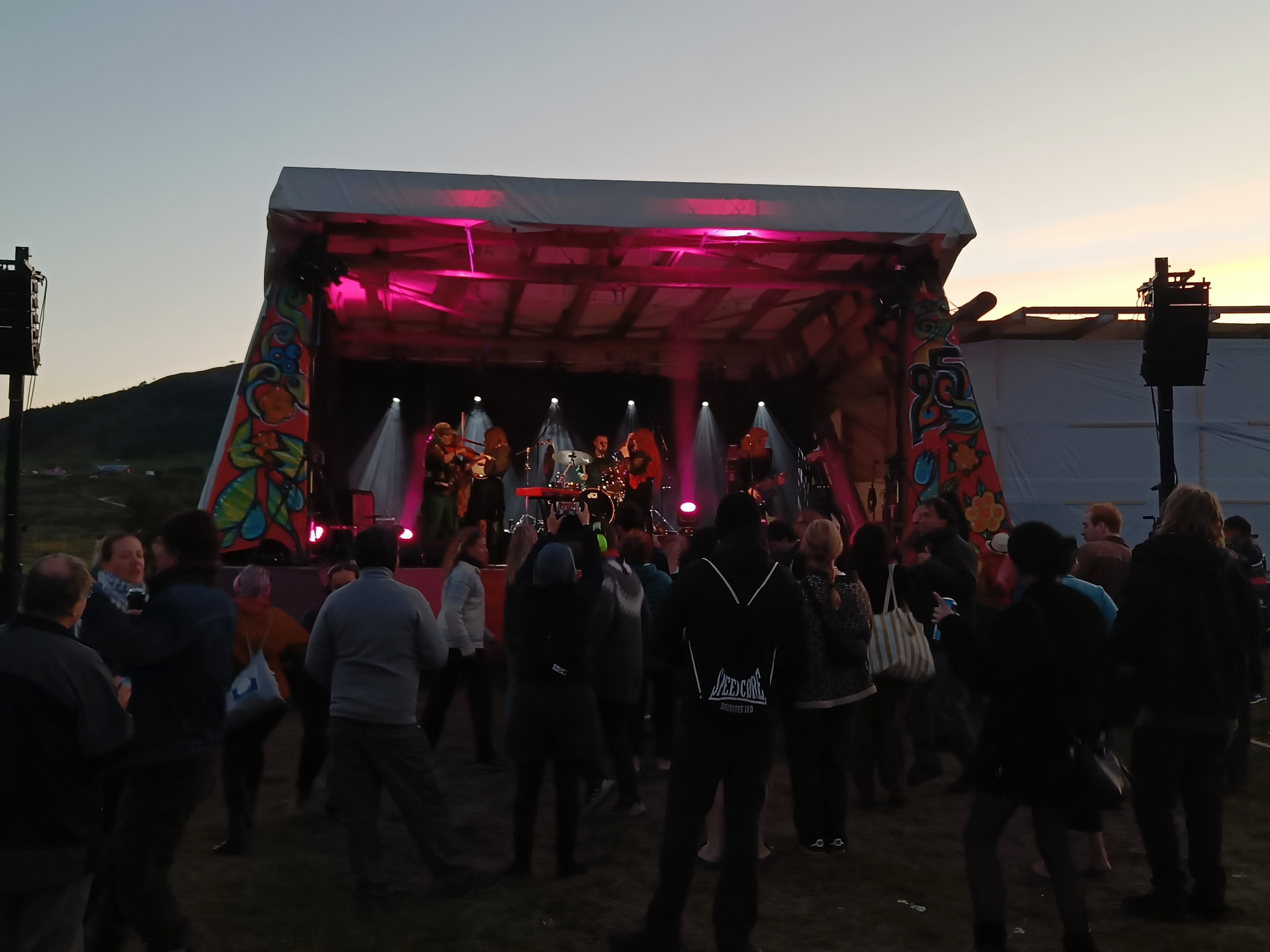 Fotografi. Karlsøyfestivalen. Bakerst i bildet er det et åpent landskap, i forgrunnen er det en fargerik utescene med et band som spiller. Fremst i bildet er det publikummere med ryggen til som ser bandet.  