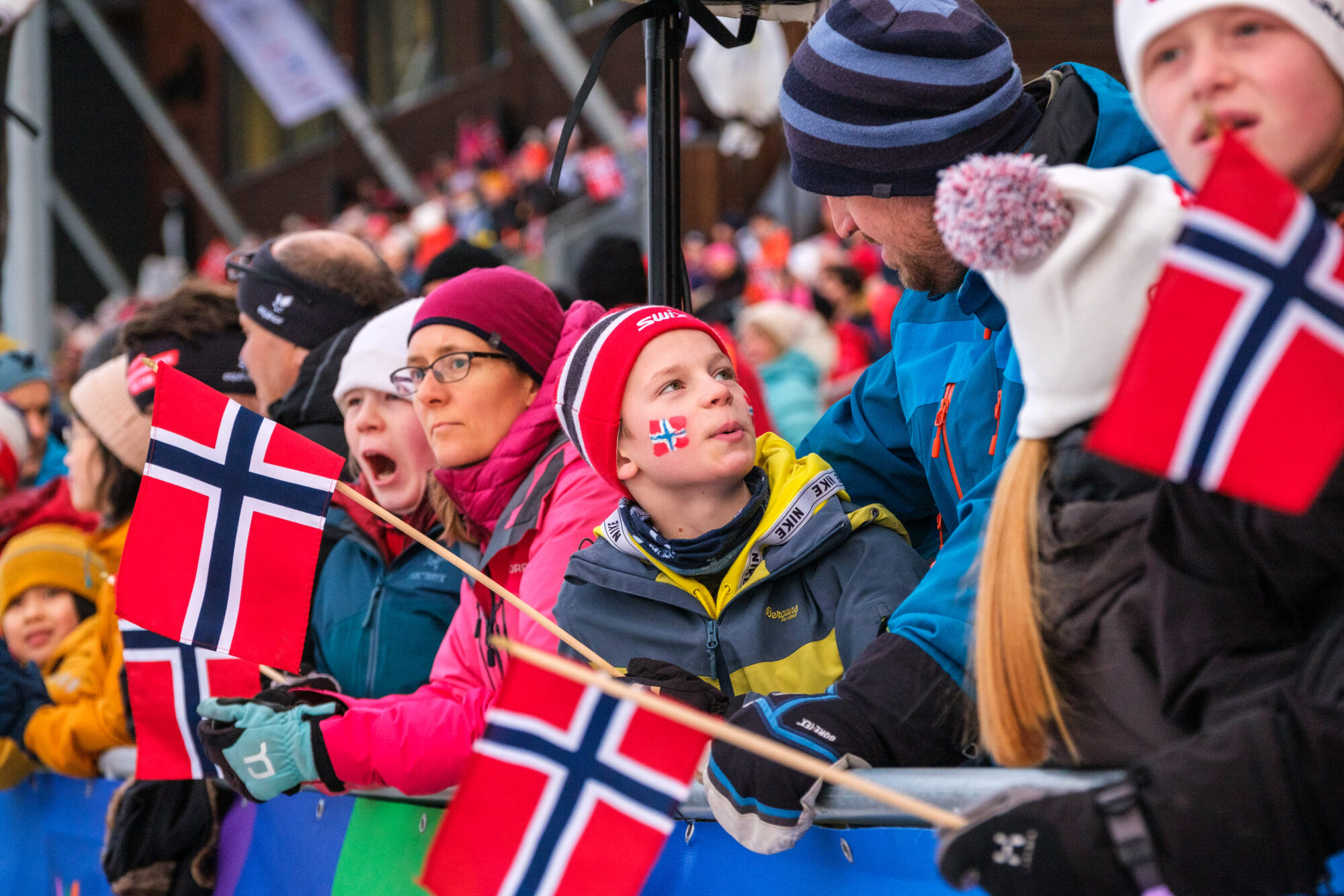 Bildet viser flere barn og voksne ved en tribune med norsk flagg og flagget malt i ansiktet, som heier på Norge.