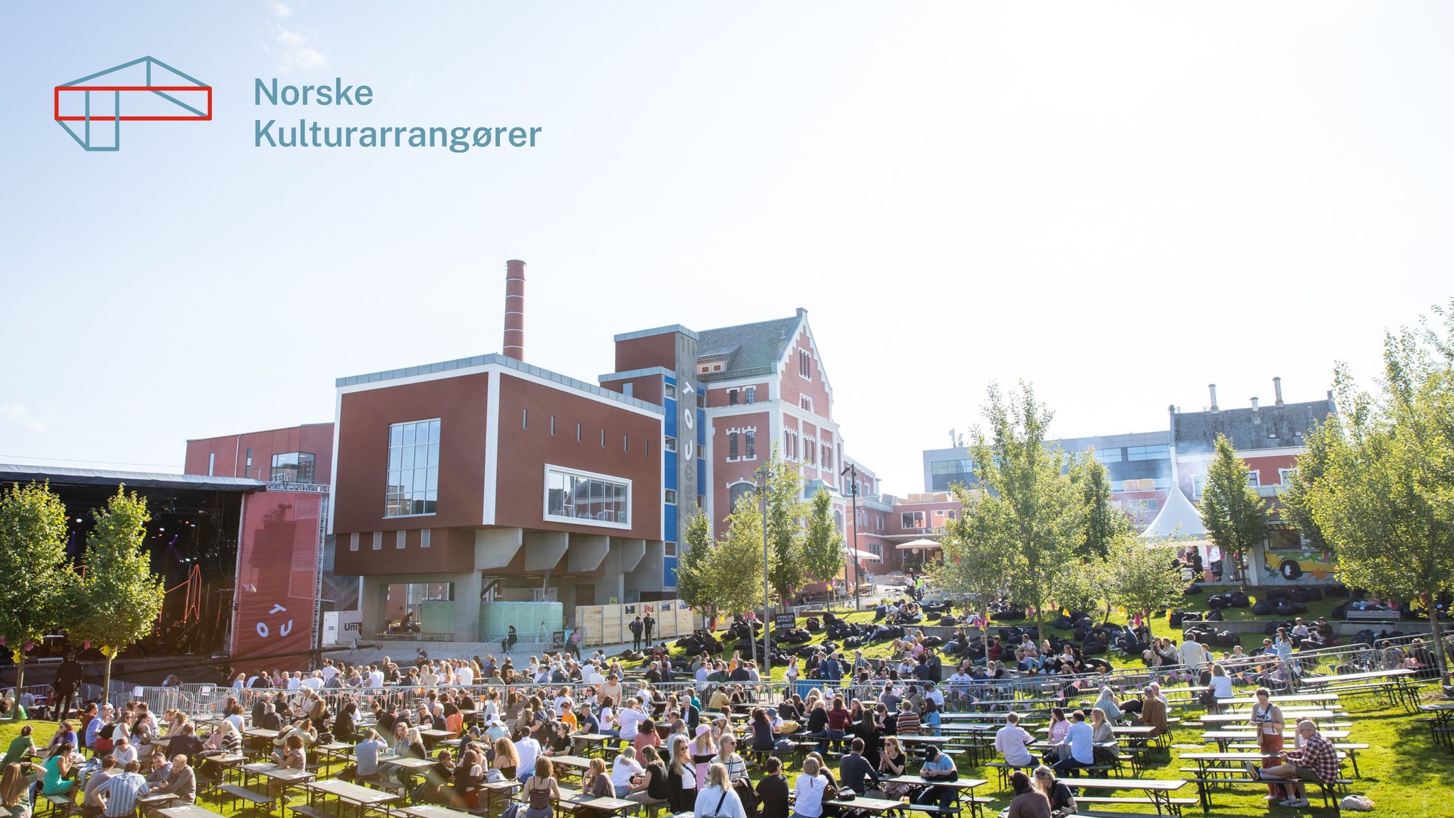 Sommerstemning på Tou. Ved siden av Tou, et gammelt industribygg, er det rigget til stor utescene og benker og bord tett i tett på en gressslette hvor folk sitter og koser seg i sola. 