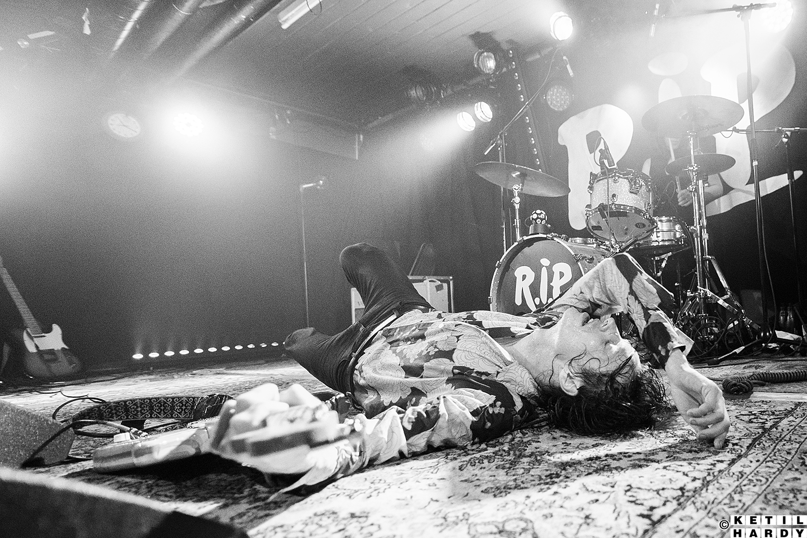 Musiker ligger på ryggen på scenen med en gitar i hånden. I bakgrunnen ses et trommesett og resten av den tomme scenen. Musikeren er tydelig utslittetter å ha spilt konsert.