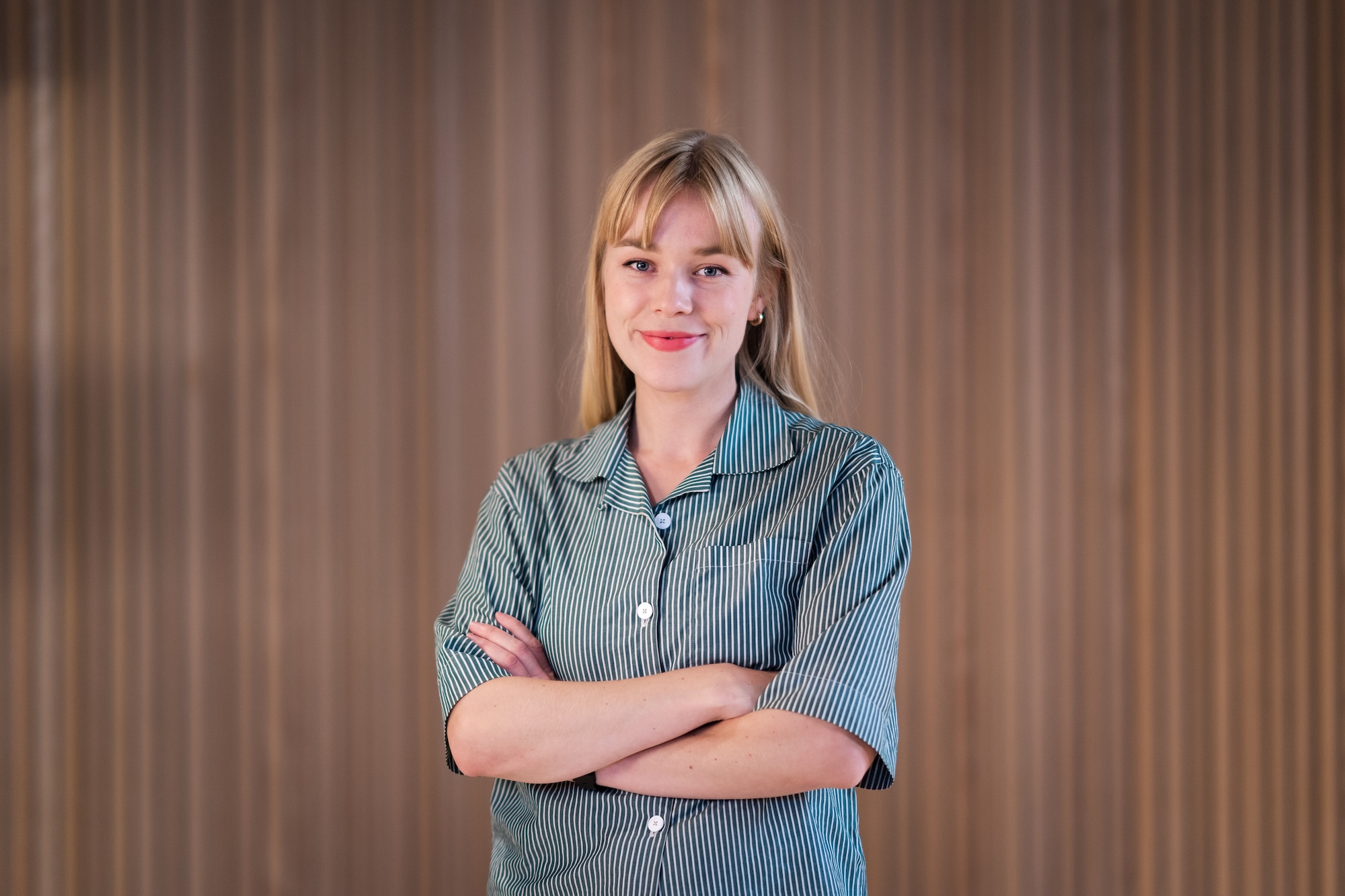 En blond kvinne smiler mot kamera med armene i kryss.