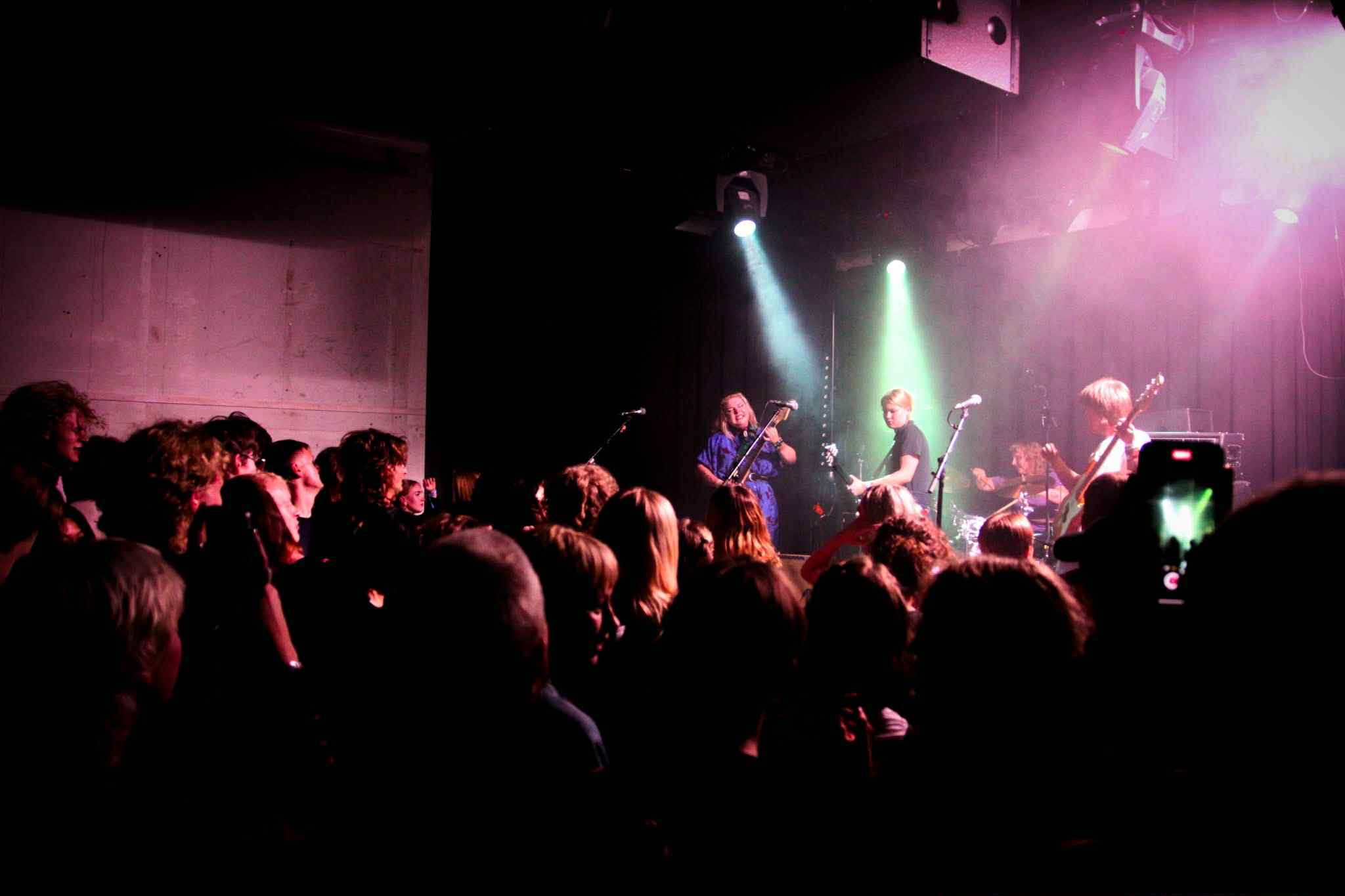 Et dunkelt lyssatt rom med et band på scenen og en folkemengde foran.