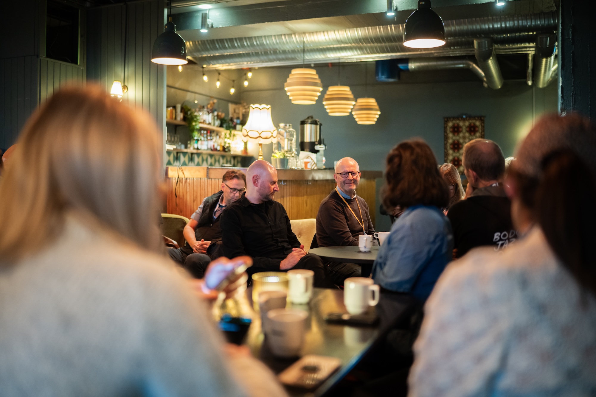 Bildet viser en gruppe mennesker som sitter iet kaféliknende rom. En smilende mann i bakgrunnen er i fokus.